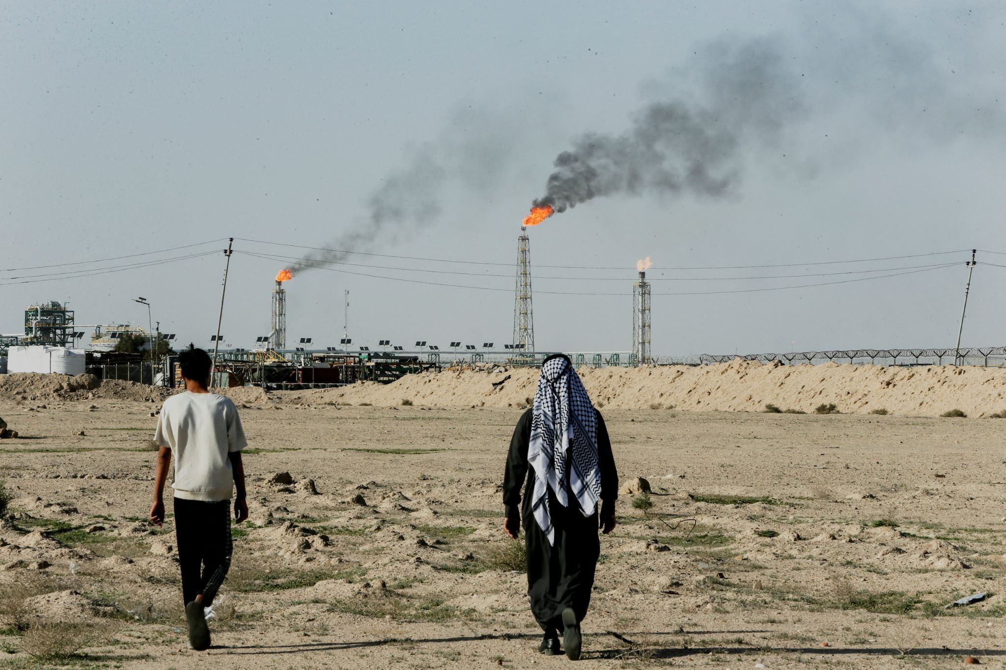 People walk near farmland by the Zubair oil field as gas flares rise in the distance