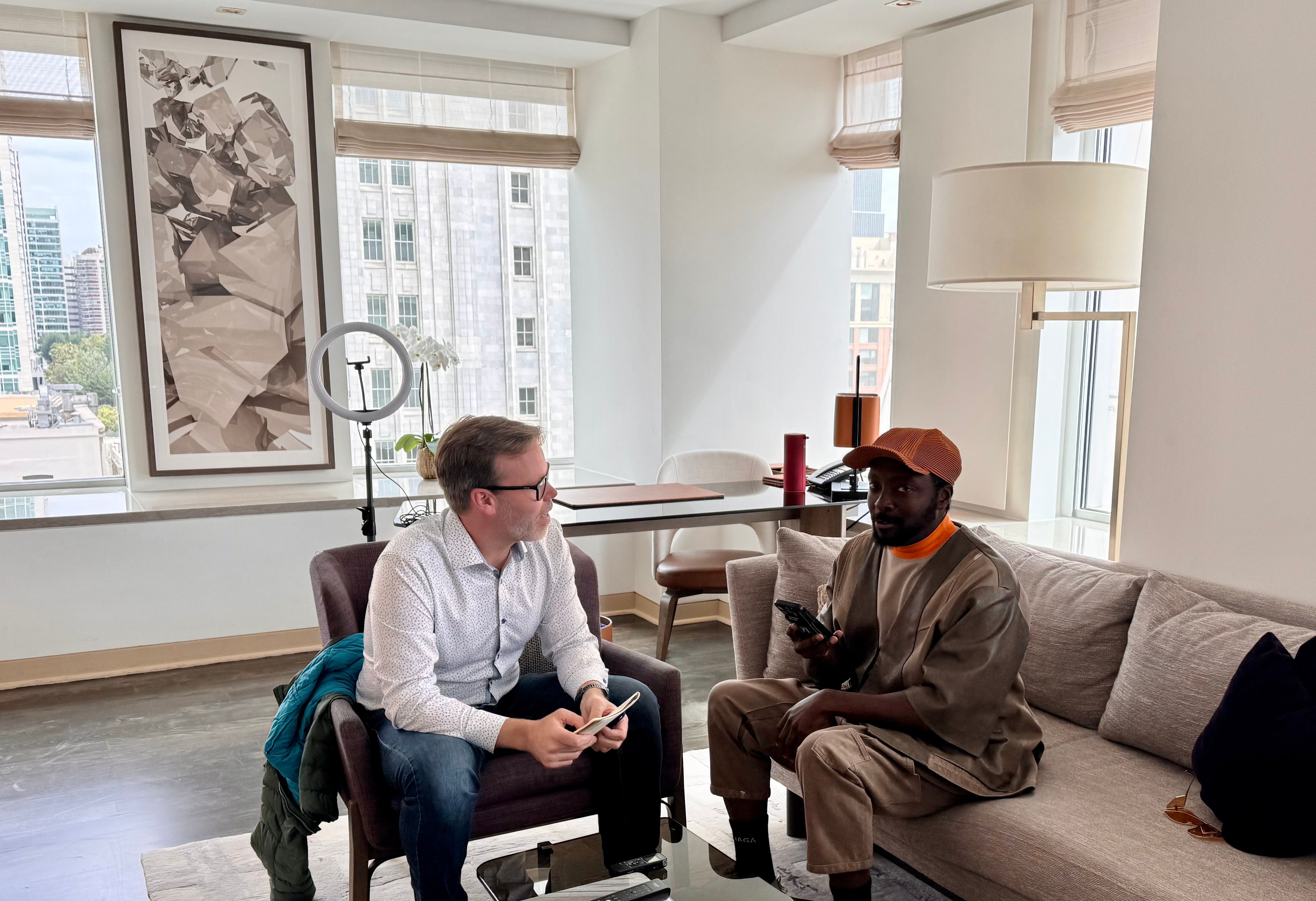 Semafor’s Reed Albergotti and singer will.i.am sitting in a living room. Buildings can be seen outside the windows.