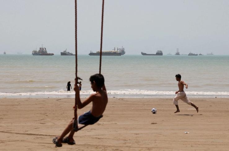 A child plays at the beach, with vessels in the Strait of Hormuz visible near the beach of Bandar Abbas