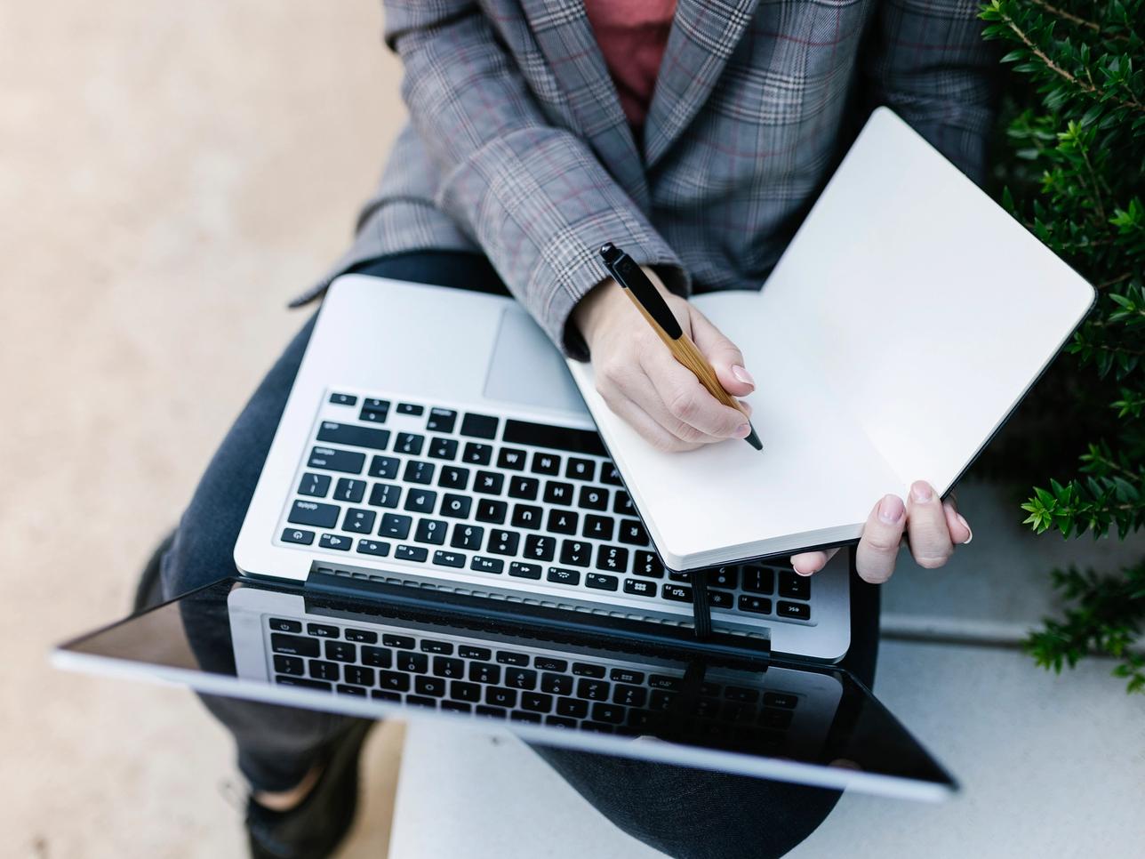 A person using a laptop and writing on a notepad.