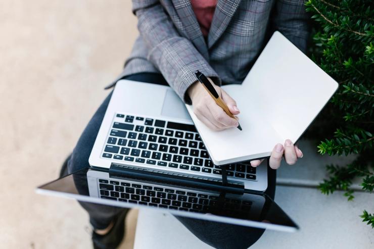 A person using a laptop and writing on a notepad.