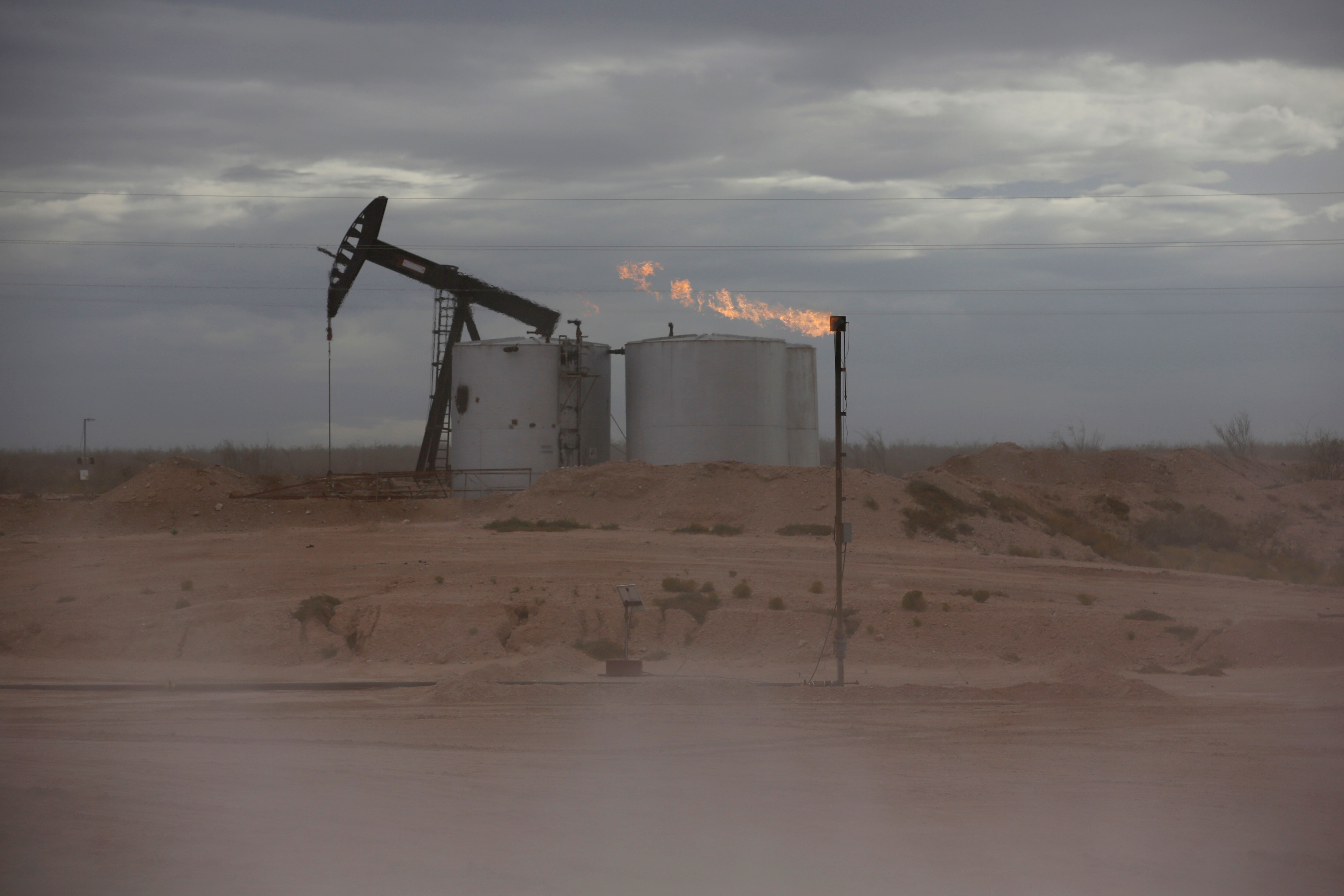 Dust blows around a crude oil pump jack and flare burning excess gas at a drill pad.
