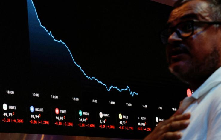A person stands near an electronic board displaying information on recent fluctuations of market indices at the B3 Stock Exchange.