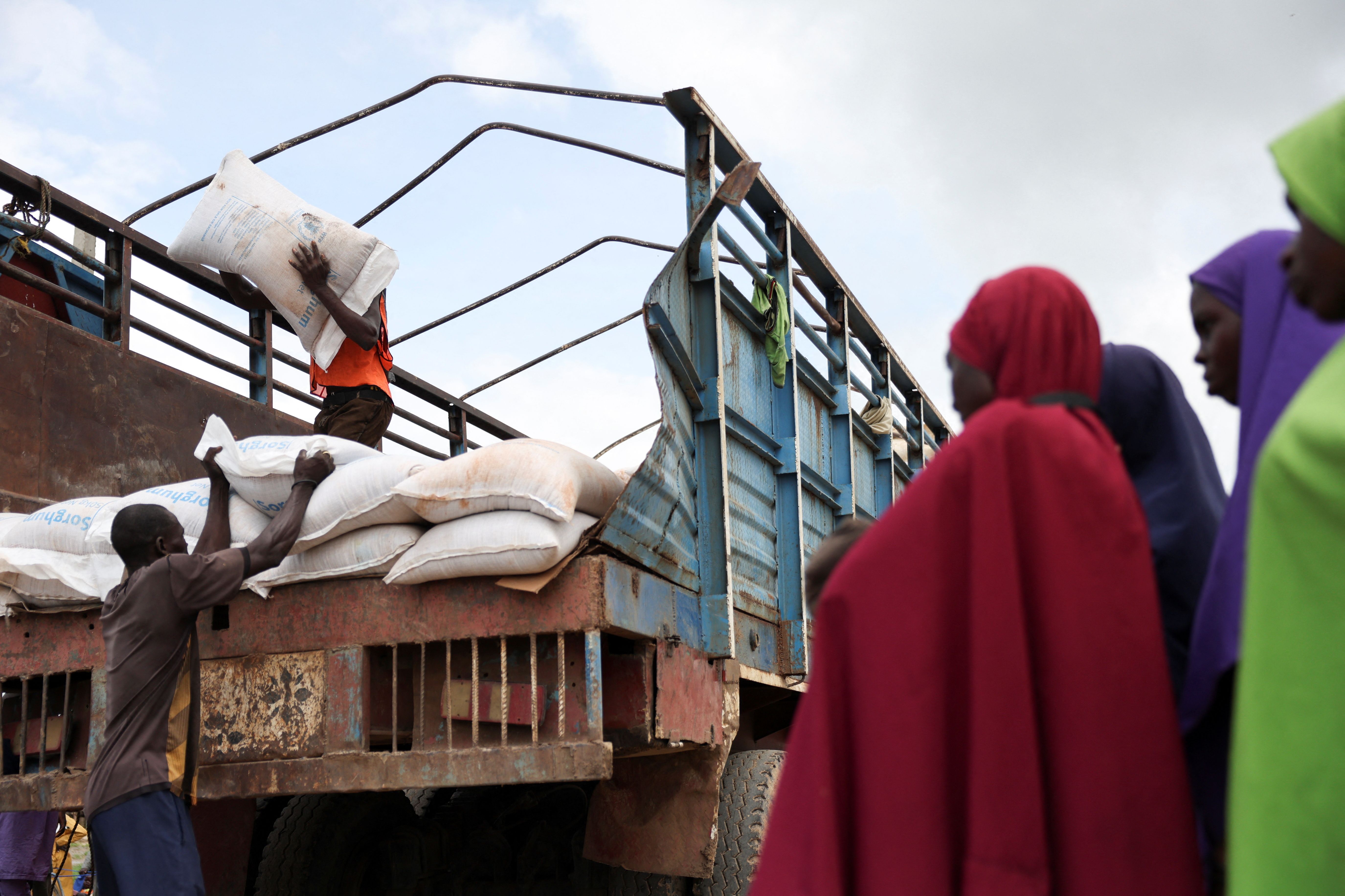Women from different Internally Displaced Persons camps look on as grains are offloaded from a truck into an open field.