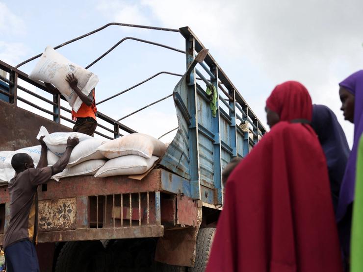 Women from different Internally Displaced Persons camps look on as grains are offloaded from a truck into an open field.