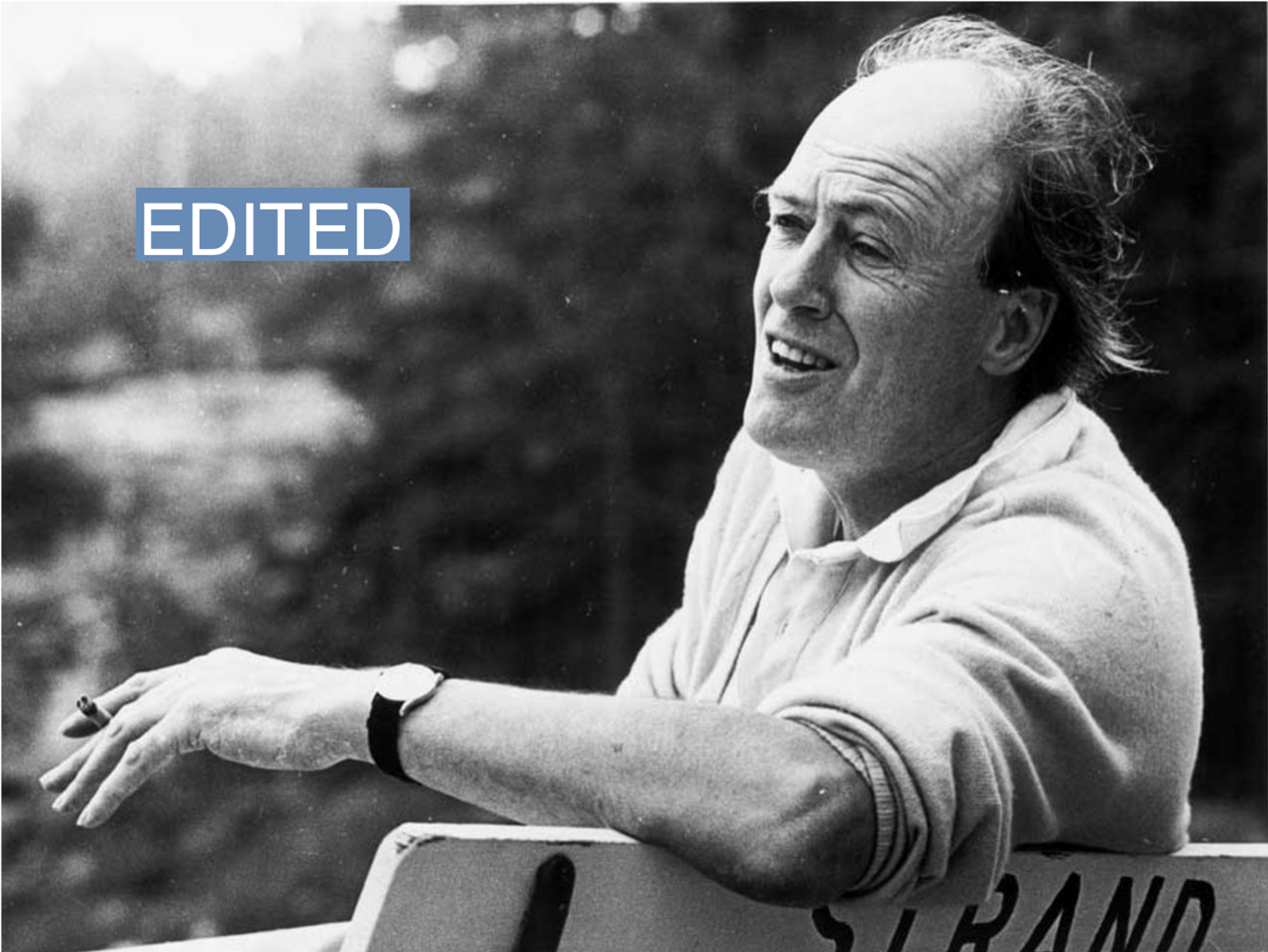 A black and white photo of Roald Dahl shows him smoking a cigarette on a bench.