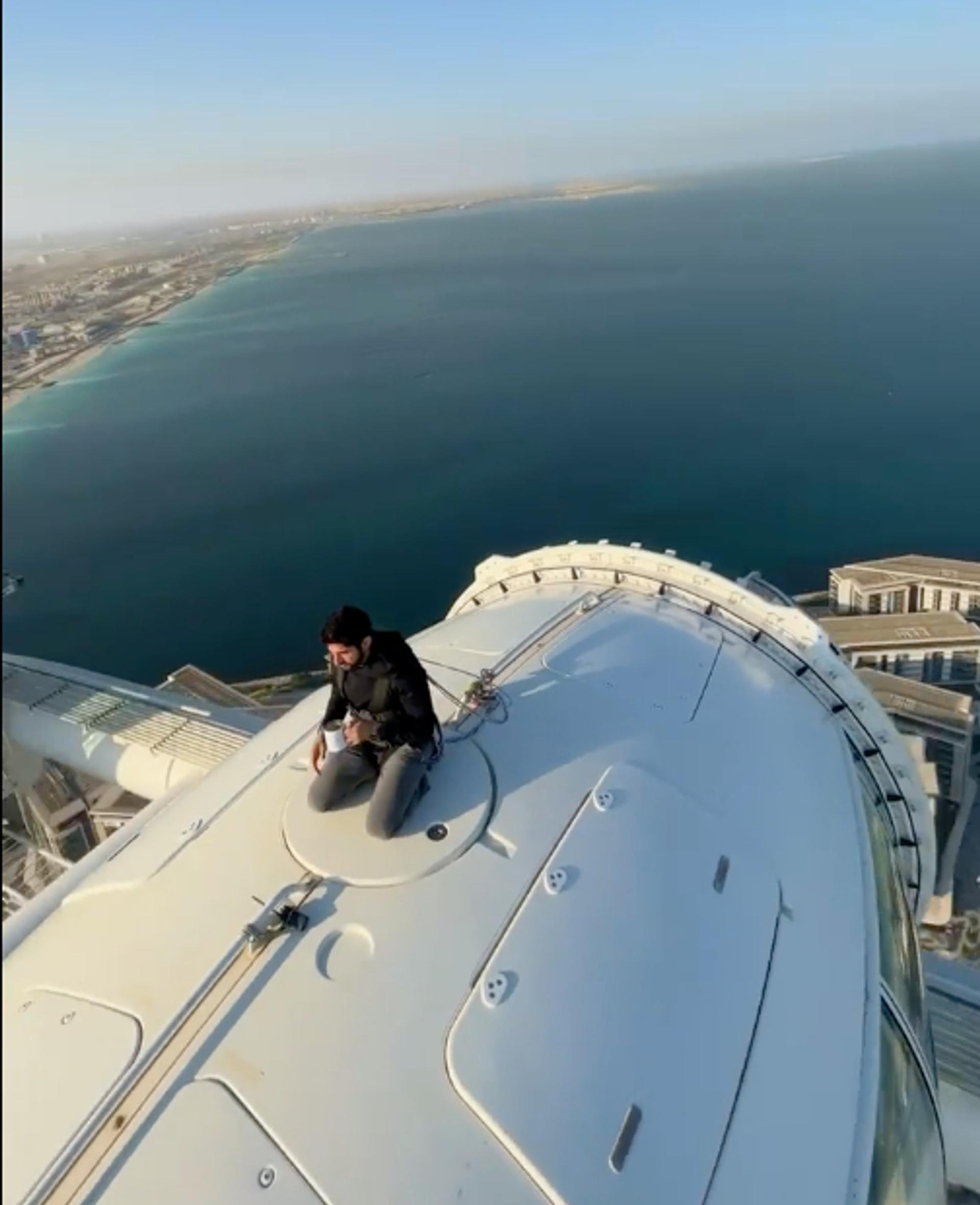 A man sits atop on a Ferris wheel cabin. The sea and sky can be seen in the background.