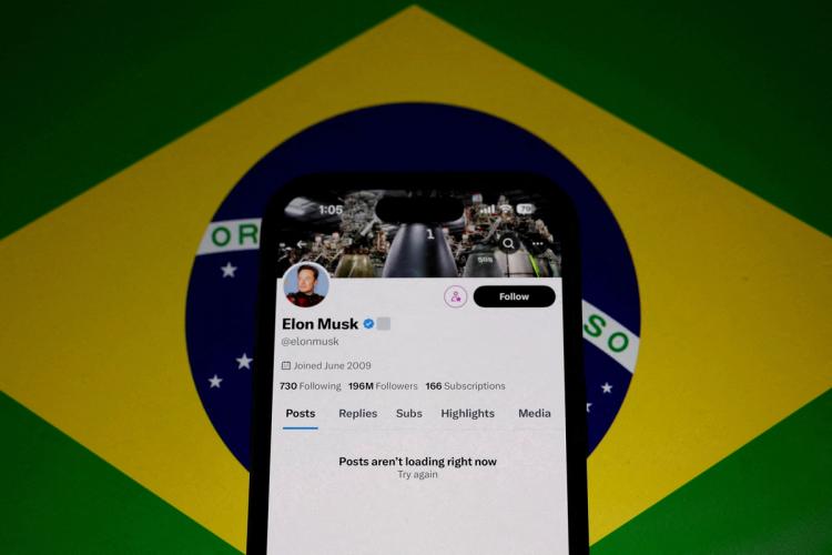 La cuenta de Elon Musk en la red social X (anteriormente Twitter) en la pantalla de un teléfono móvil, con un mensaje que indica que las publicaciones no se están cargando en ese momento. El fondo está compuesto por una bandera de Brasil, lo que sugiere una conexión con la reciente suspensión de la plataforma en el país.