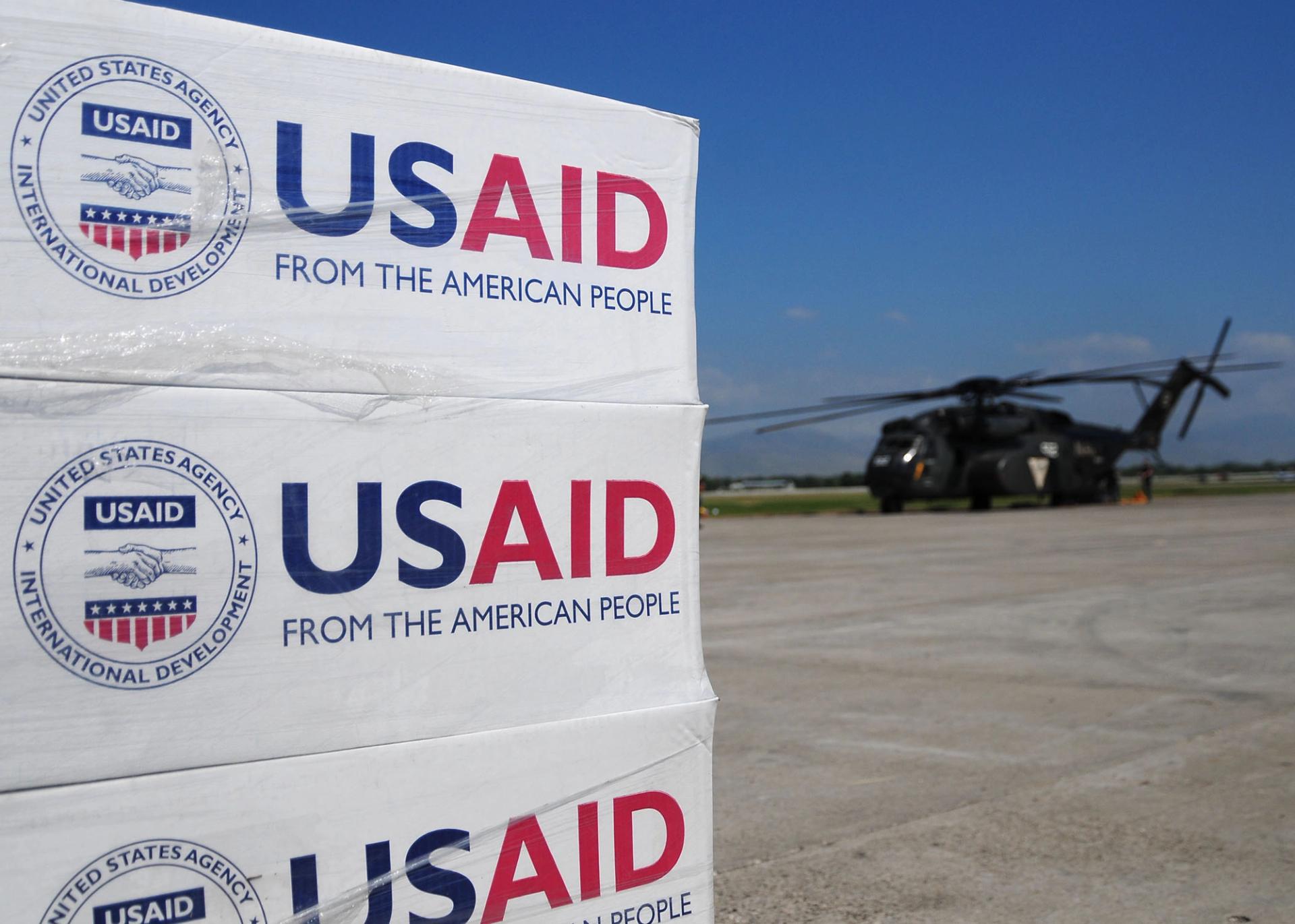 Pallets of USAID supplied set to be delivered with a helicopter in the background.