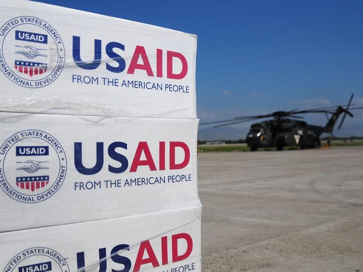 Pallets of USAID supplied set to be delivered with a helicopter in the background.