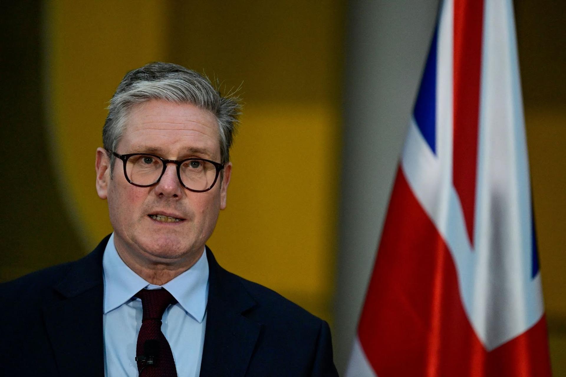 Keir Starmer, wearing a dark suit and tie, stands in front of a British flag as he addresses a press conference in Berlin