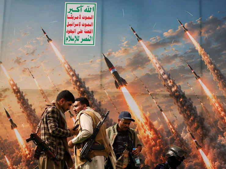 Two Houthi supporters stand in front of a picture of missiles in the sky.