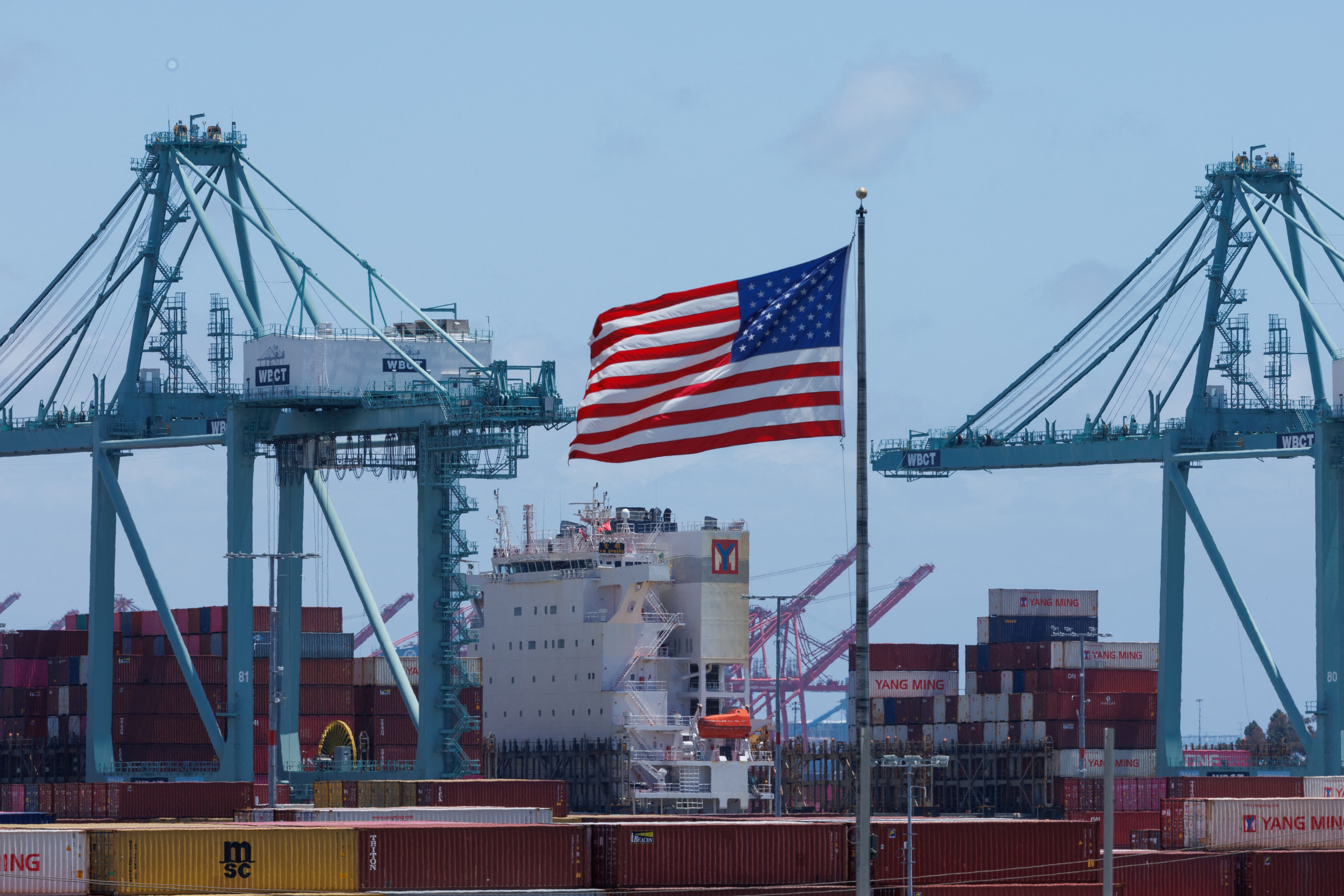 An American flag flutters over a ship and shipping containers at the Port of Los Angeles