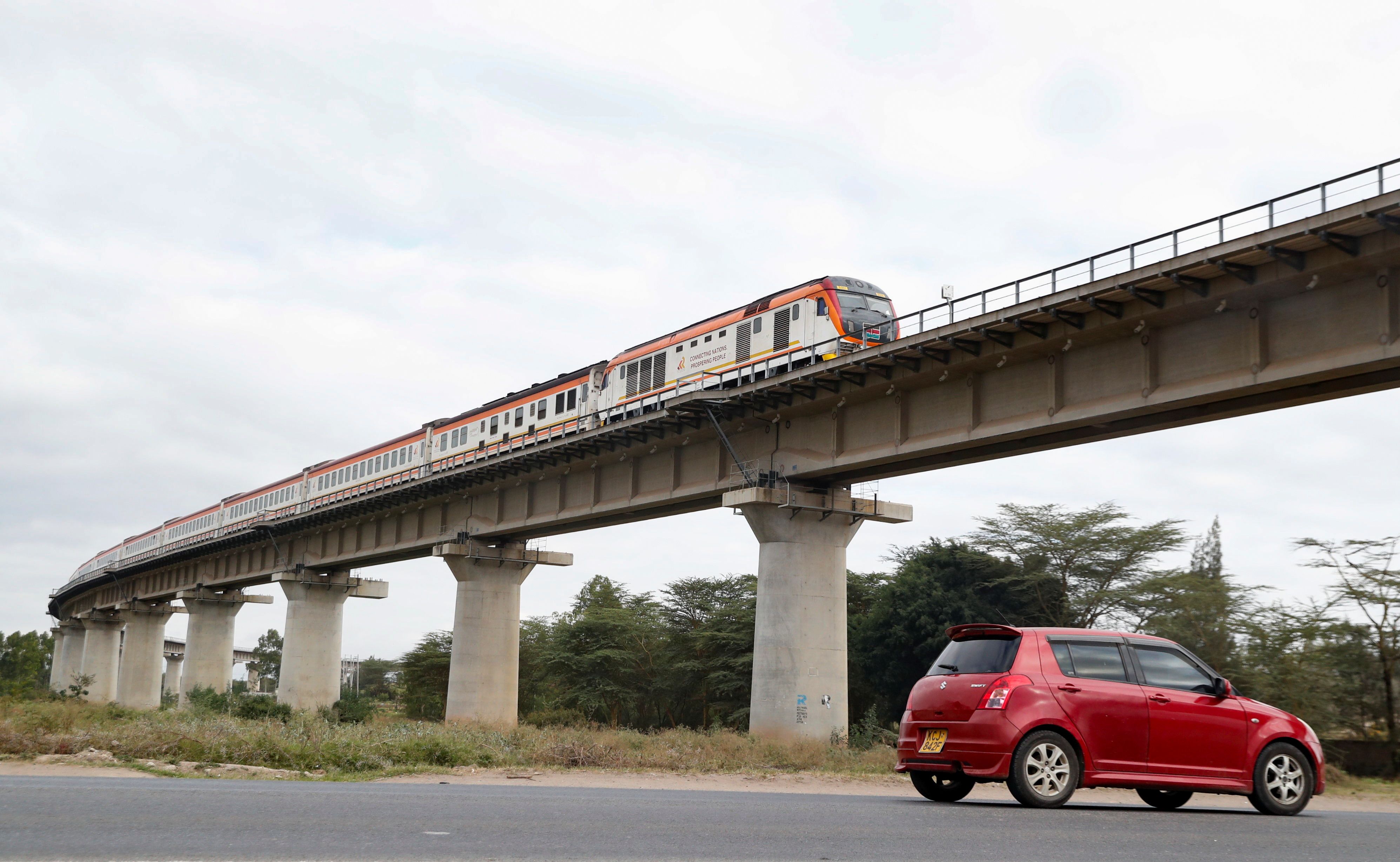 A car passes under a bridge as a train moves along the Standard Gauge Railway.