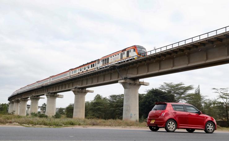 A car passes under a bridge as a train moves along the Standard Gauge Railway.