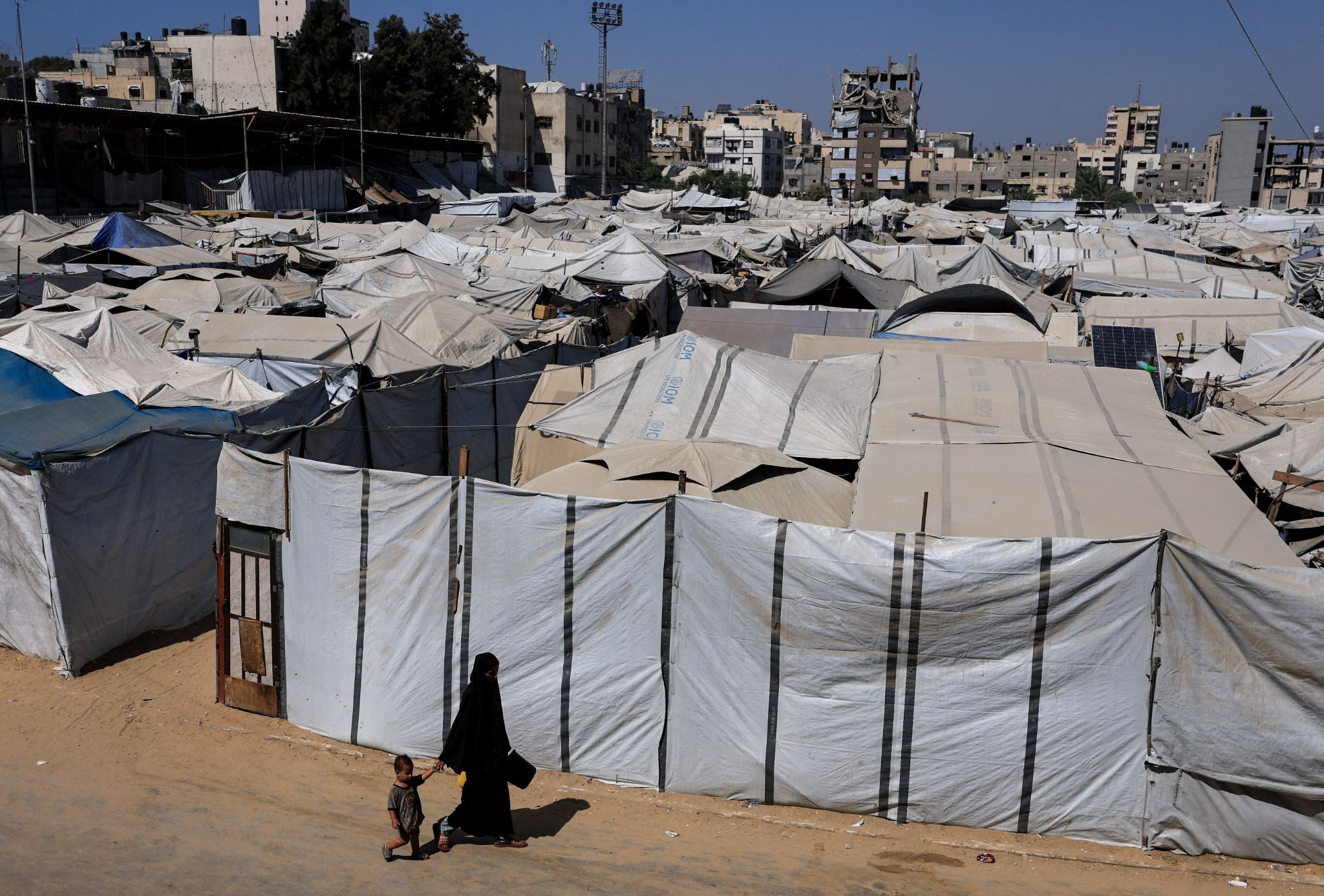 A Palestinian woman and a child walk next to a tent camp sheltering displaced Palestinians in Gaza City. 