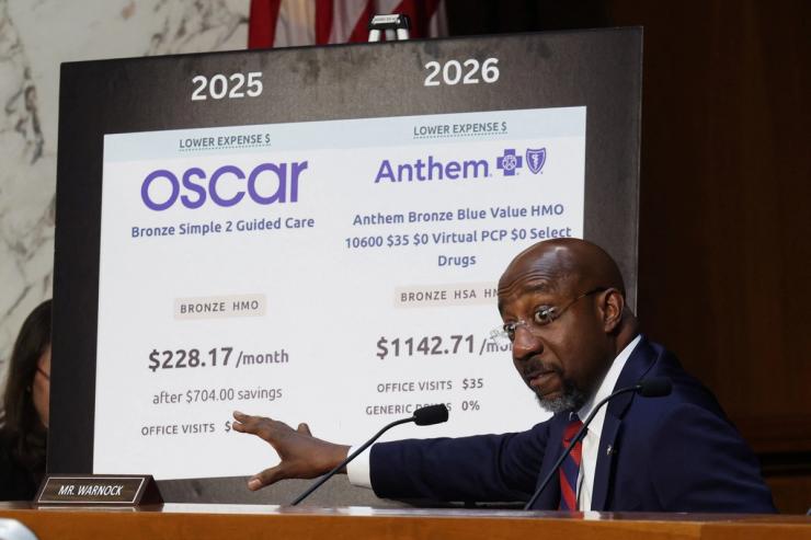 Raphael Warnock gestures to a sign featuring health insurance information