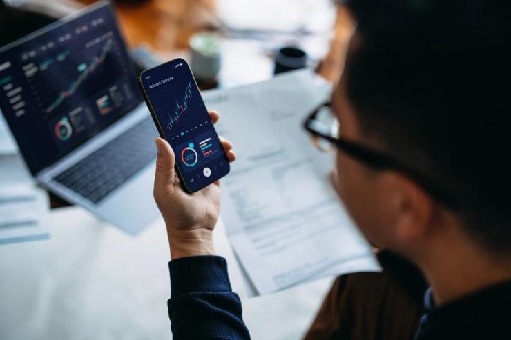 Young Asian man managing finance and investment online, analyzing stock market trades with smartphone and laptop.