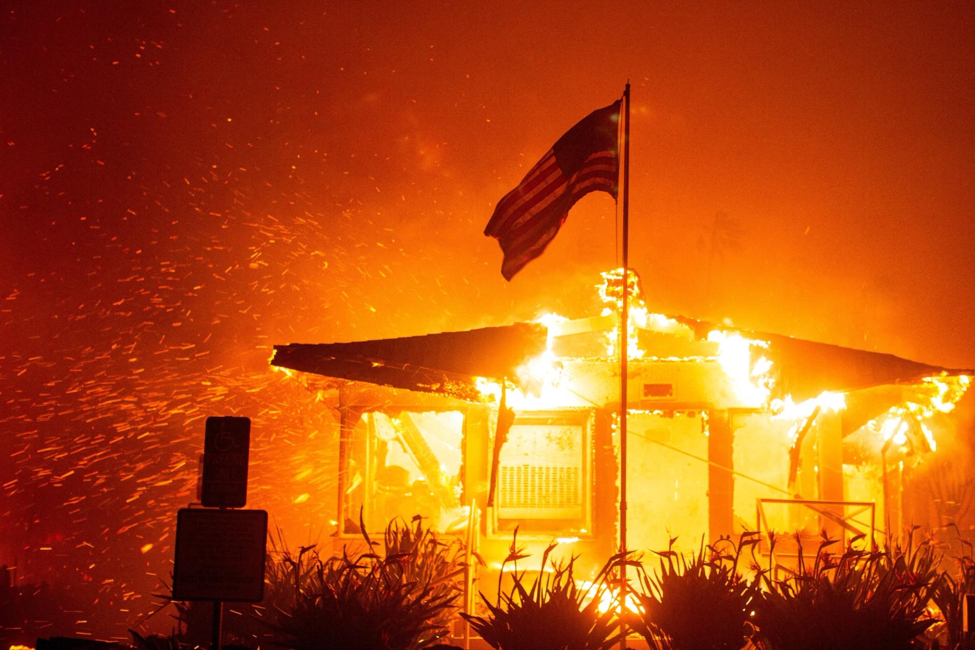 A U.S flag flies as fire engulfs a structure while the Palisades Fire burns during a windstorm on the west side of Los Angeles, California, U.S. January 7, 2025.