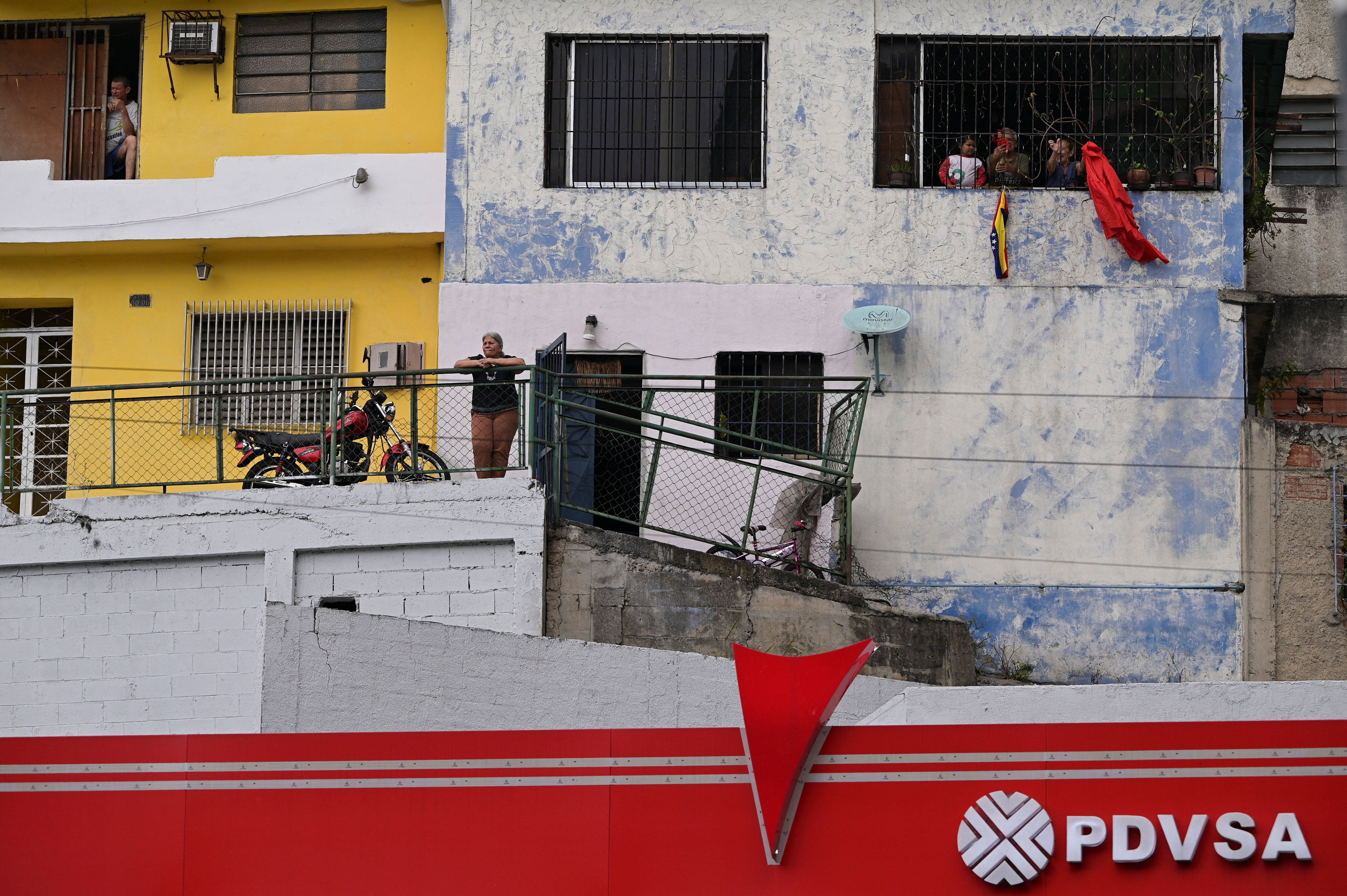 The logo of Venezuelan state oil company PDVSA is seen as a Venezuelan flags hangs from a window while people watch a march of government supporters calling for the release of Venezuela’s ousted president.