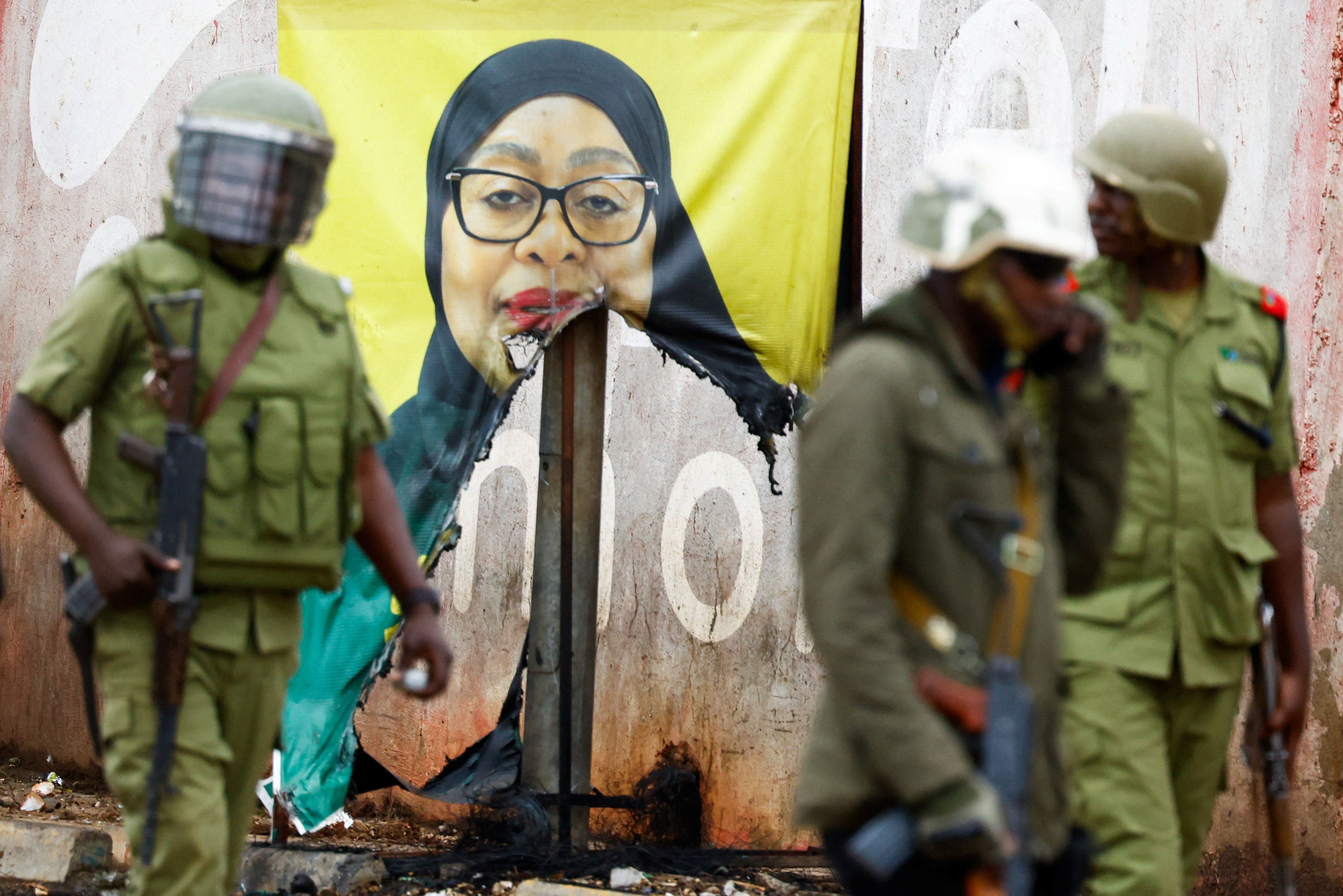 Tanzanian riot police officers walk past a vandalised campaign poster of President Samia Suluhu Hassan.