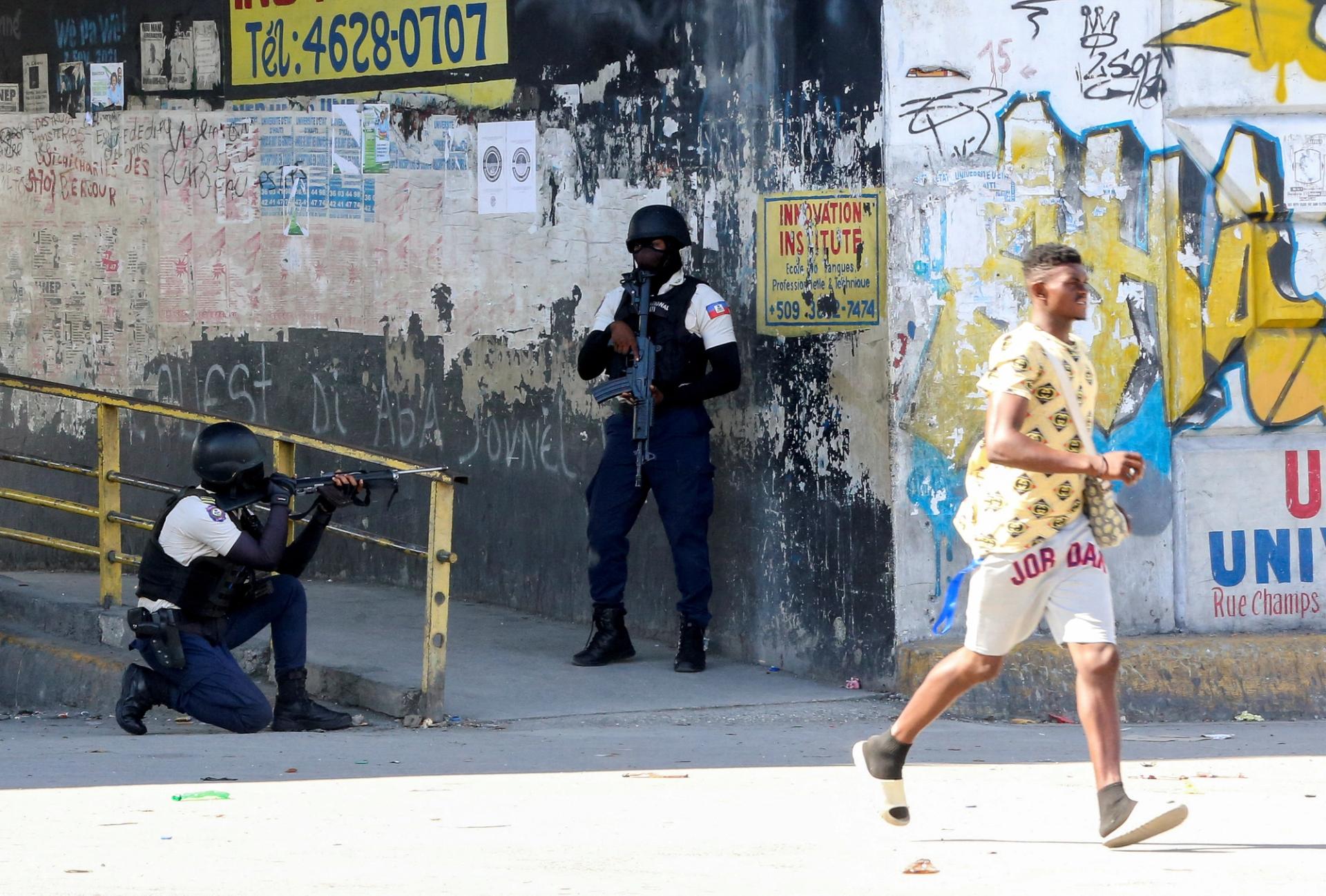 A photo of two members of Haiti’s security forces taking cover while a man runs past them. 