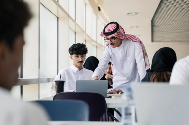 University classmates sitting at tables, doing homework, completing assignments, interacting with mature educator.