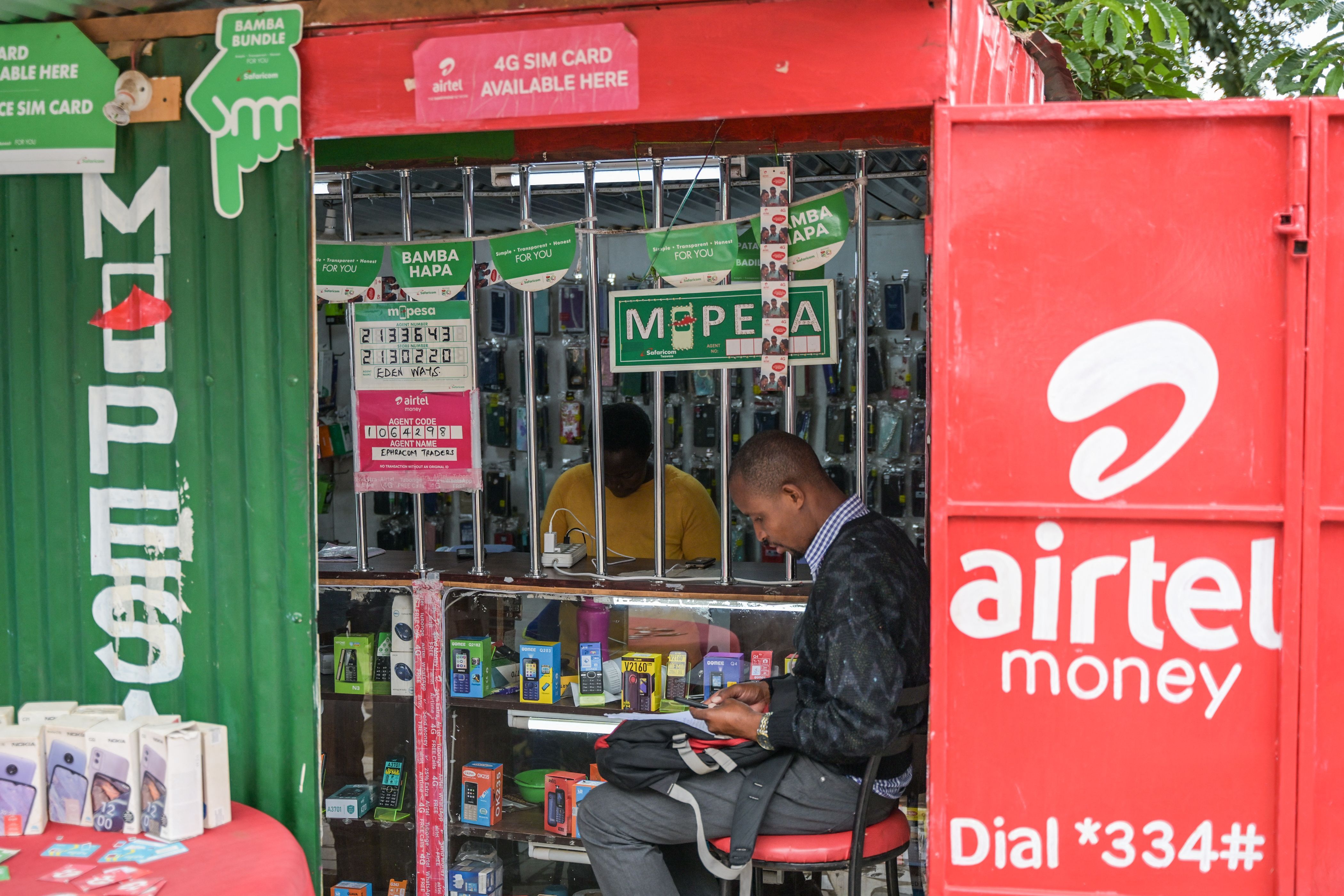 A customer sits at a kiosk next to a sign advertising M-Pesa, Safaricom’s and Airtel mobile money service, in Nairobi, on September 14, 2023. 