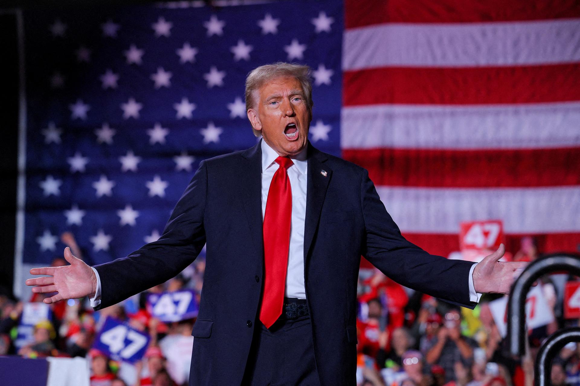 Donald Trump, wearing a navy suit and red tie, pictured with his hands outspread at a rally in Michigan